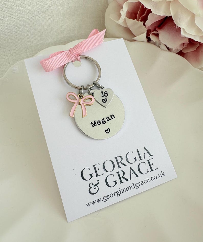 May include: A silver keychain with a pink bow and a charm that says "Megan" with a heart symbol. The keychain also has a heart-shaped charm with the number "18" and a pink ribbon. The card below the keychain has the text "GEORGIA & GRACE".