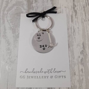 May include: Silver keychain with a round charm that says "Ben" with a star on each side. The charm is attached to a smaller charm that says "21" with a star. A silver hammer charm is also attached to the keychain. The keychain is on a white card with the text "handmade with love" and "GG JEWELLERY & GIFTS".