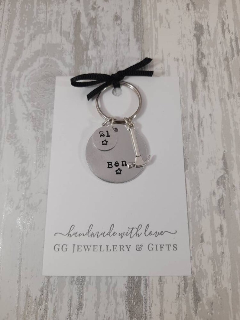 May include: Silver keychain with a round charm that says "Ben" with a star on each side. The charm is attached to a smaller charm that says "21" with a star. A silver hammer charm is also attached to the keychain. The keychain is on a white card with the text "handmade with love" and "GG JEWELLERY & GIFTS".