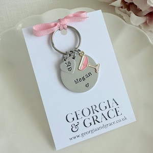 May include: A silver-colored keychain with a pink ribbon, a round charm engraved with "Megan" and a heart, and a charm shaped like a cocktail glass. The keychain also has a heart-shaped charm with the number "18". The card reads "GEORGIA & GRACE".