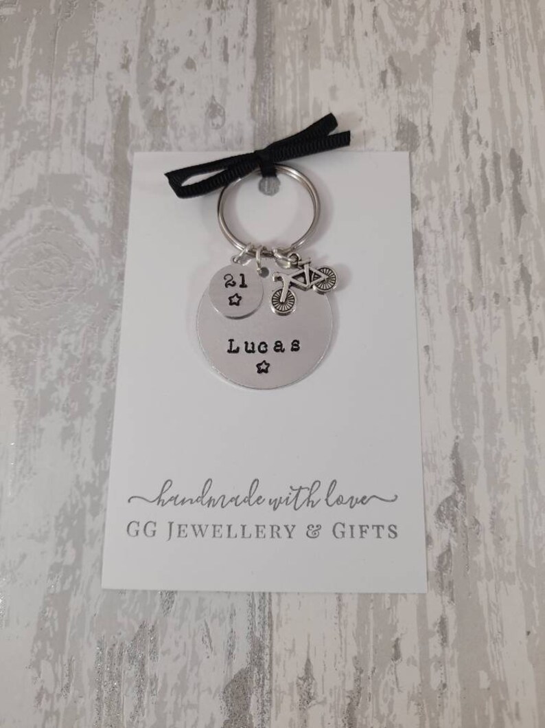 May include: Silver keychain with a round charm that says "Lucas" and a smaller charm with the number "21" and a star. The keychain also has a small charm of a bicycle. The card attached to the keychain says "handmade with love GG JEWELLERY & GIFTS".