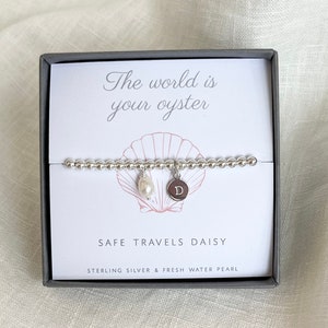 K&ouml;nnte beinhalten: Ein silbernes Armband mit einem Perlenanh&auml;nger und einem silbernen Anh&auml;nger mit dem Buchstaben "D" darauf. Das Armband befindet sich in einer Schachtel mit dem Text "Die Welt ist deine Auster" und "Safe Travels Daisy" darauf.