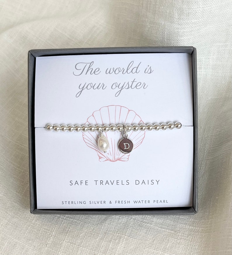 K&ouml;nnte beinhalten: Ein silbernes Armband mit einem Perlenanh&auml;nger und einem silbernen Anh&auml;nger mit dem Buchstaben "D" darauf. Das Armband befindet sich in einer Schachtel mit dem Text "Die Welt ist deine Auster" und "Safe Travels Daisy" darauf.