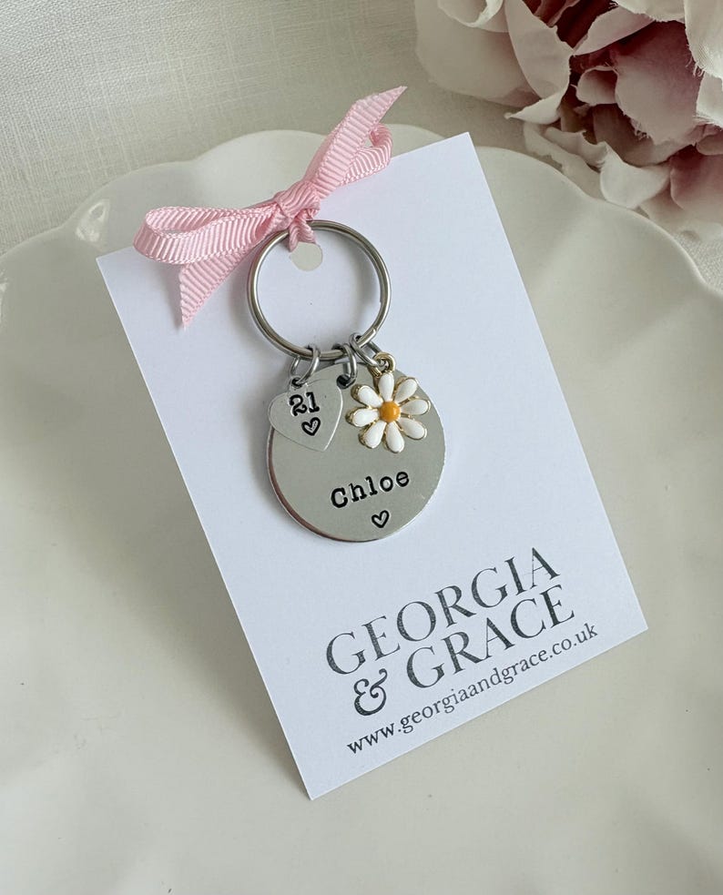 Pu&ograve; includere: Un portachiavi d'argento con un ciondolo a forma di margherita, un ciondolo a forma di cuore con il numero 21 e un'etichetta rotonda che dice "Chloe" con un cuore. Il portachiavi &egrave; su una carta bianca con il testo "GEORGIA & GRACE www.georgiaandgrace.co.uk"