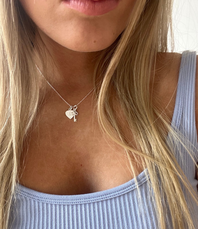 Op de afbeelding: Een zilveren ketting met een hartvormige hanger en een sleutel hanger. De hartvormige hanger heeft een kleine inscriptie.