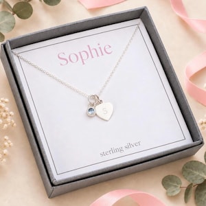 May include: A silver necklace with a small heart-shaped charm engraved with the letter S and a round blue crystal charm. The necklace is displayed on a white card inside a dark gray box, with the name Sophie printed on the card.