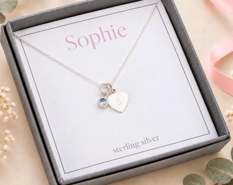 Op de afbeelding: Een zilveren ketting met een kleine hartvormige bedel gegraveerd met de letter S en een ronde blauwe kristallen bedel. De ketting wordt gepresenteerd op een witte kaart in een donkergrijze doos, met de naam Sophie op de kaart gedrukt.