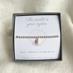 K&ouml;nnte beinhalten: Ein silbernes Armband mit einem S&uuml;&szlig;wasserperlen-Anh&auml;nger in einer grauen Schachtel. Die Schachtel enth&auml;lt eine wei&szlig;e Karte mit dem Text "The world is your oyster" und "Safe Travels Charlotte". Die Karte tr&auml;gt au&szlig;erdem den Hinweis "Sterling Silver & Fresh Water Pearl".