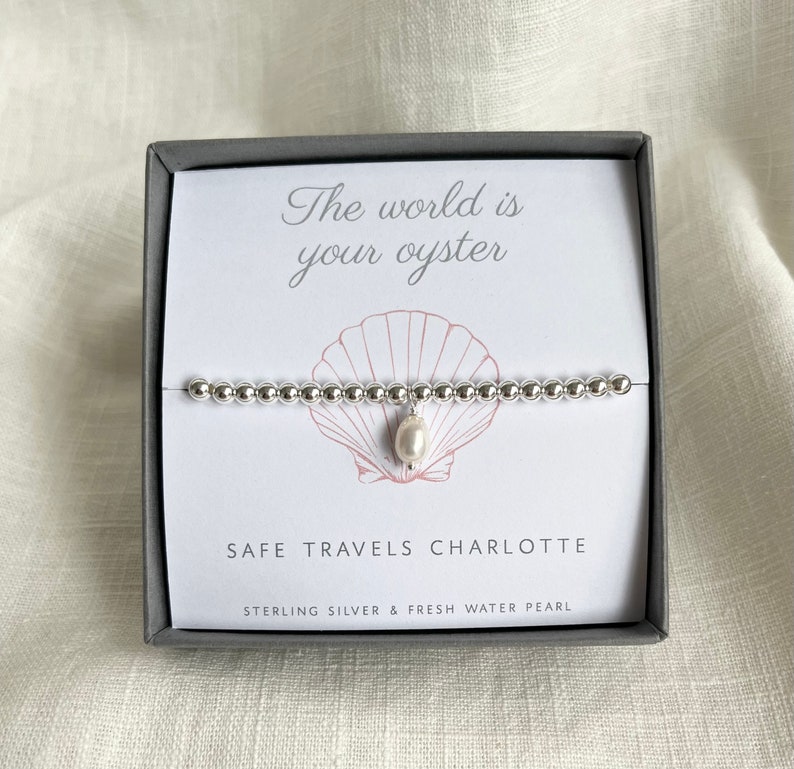 K&ouml;nnte beinhalten: Ein silbernes Armband mit einem S&uuml;&szlig;wasserperlen-Anh&auml;nger in einer grauen Schachtel. Die Schachtel enth&auml;lt eine wei&szlig;e Karte mit dem Text "The world is your oyster" und "Safe Travels Charlotte". Die Karte tr&auml;gt au&szlig;erdem den Hinweis "Sterling Silver & Fresh Water Pearl".