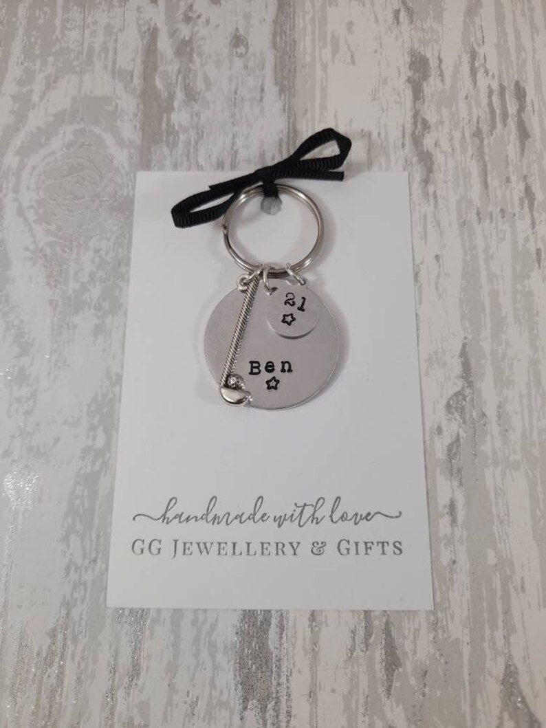 May include: A silver keychain with a golf club charm and a round tag that says "Ben" with a star. A smaller round tag with the number "21" and a star is attached to the keychain. The keychain is on a white card with the text "handmade with love" and "GG JEWELLERY & GIFTS".