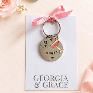 May include: A silver-toned metal keychain featuring a round disc stamped with the name Megan and a small heart, a smaller disc with the number 21 and a heart, and a cocktail glass charm. The keychain is attached to a white card labeled GEORGIA & GRACE.