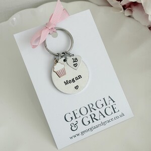 May include: A silver keychain with a cupcake charm, a heart charm with the number 18, and a heart charm. The keychain is attached to a white card with the name "Megan" and the text "GEORGIA & GRACE". A pink ribbon is tied to the keychain.
