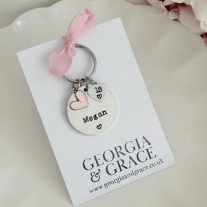 May include: A personalized silver keychain with a pink ribbon. The keychain features a round charm engraved with the name "Megan" and a heart symbol, plus two heart-shaped charms, one with a pink heart and the other with the number "18".