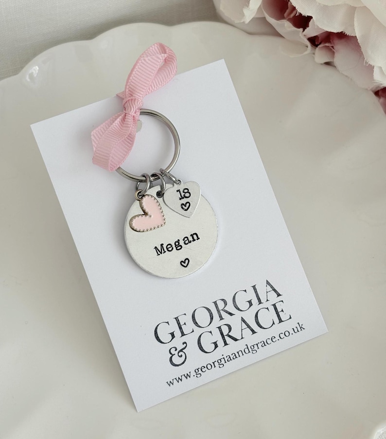 May include: A personalized silver keychain with a pink ribbon. The keychain features a round charm engraved with the name "Megan" and a heart symbol, plus two heart-shaped charms, one with a pink heart and the other with the number "18".