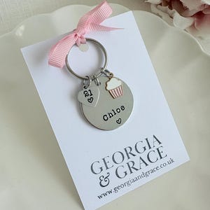 Pu&ograve; includere: Un portachiavi argentato con un ciondolo a forma di cupcake, un ciondolo a forma di cuore con il numero 21 e un'etichetta rotonda che dice "Chloe". Il portachiavi &egrave; su una carta bianca con il testo "GEORGIA & GRACE www.georgiaandgrace.co.uk"