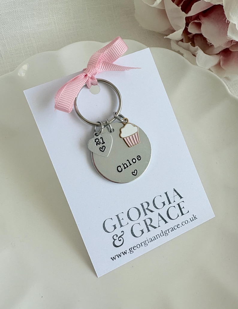 Pu&ograve; includere: Un portachiavi argentato con un ciondolo a forma di cupcake, un ciondolo a forma di cuore con il numero 21 e un'etichetta rotonda che dice "Chloe". Il portachiavi &egrave; su una carta bianca con il testo "GEORGIA & GRACE www.georgiaandgrace.co.uk"