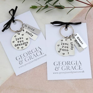May include: Two silver keychains with charms. One keychain has a round charm that says "love you Grandma" and two rectangular charms that say "Toby" and "Molly". The other keychain has a round charm that says "love you Nann" and two rectangular charms that say "Lilly" and "Grace Harry". Both keychains have a black ribbon and a white card with the text "GEORGIA & GRACE www.georgiaandgrace.co.uk"