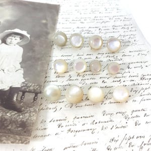 May include: A collection of iridescent, round, pearl-like buttons. The buttons are arranged on a vintage handwritten letter, with a sepia-toned photograph in the background. The buttons vary slightly in size and color.