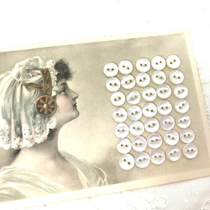 May include: A vintage postcard featuring a grayscale portrait of a woman in profile, adorned with a decorative headpiece. A collection of small, round, white buttons with two holes each are arranged on the postcard, suggesting a craft or sewing theme.