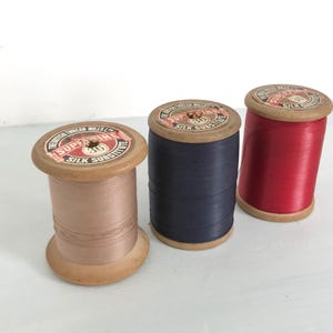 May include: Three spools of thread, each with a wooden base and a paper label. The labels are red, white, and blue and say "The Superior Thread Mills Ltd. Supreme Silk Substitute". The thread colors are beige, navy blue, and red.