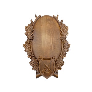 May include: A wooden plaque with a shield design featuring crossed rifles and a hunting scene. The plaque is surrounded by an oak leaf and acorn wreath and antler points.