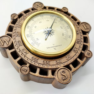 Puede incluir: Una brújula de madera con una esfera de termómetro de color dorado. La brújula tiene un diseño circular con un marco de madera y un borde de color dorado. La esfera del termómetro tiene un fondo blanco con marcas negras y una aguja negra. La brújula está marcada con las letras N, S, E y W.