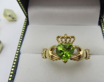 May include: A gold Claddagh ring with a heart-shaped peridot gemstone. The ring features hands holding the heart, a crown above, and small accent stones. Presented in a black jewelry box with a white interior.