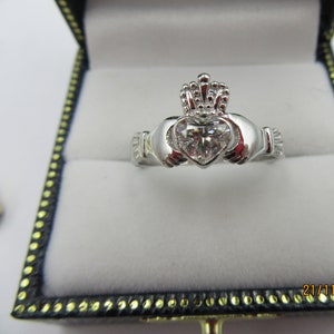 May include: A silver Claddagh ring with a heart-shaped diamond in the center and a crown on top. The ring is in a white velvet ring box.