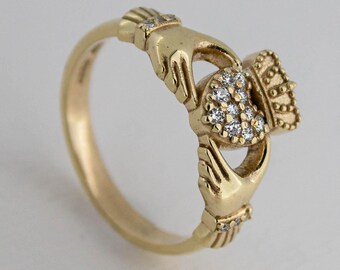 Puede incluir: Un anillo Claddagh de oro que muestra dos manos sosteniendo un corazón coronado. El corazón está incrustado con pequeñas piedras claras y brillantes, y los puños de las manos están decorados con diminutas piedras claras.