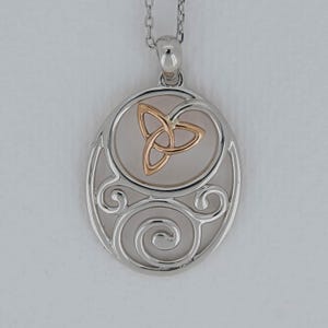 Puede incluir: Un colgante ovalado de tono plateado con un diseño calado de patrones metálicos en espiral. En la parte superior central se encuentra un símbolo de nudo triquetra en tono oro rosa. El colgante cuelga de una cadena plateada a juego.