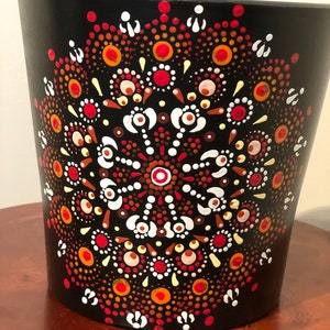 May include: Black cylindrical container with a hand-painted mandala design. The mandala features a symmetrical pattern of red, orange, white, and brown dots on a black background. The container sits on a wooden surface.