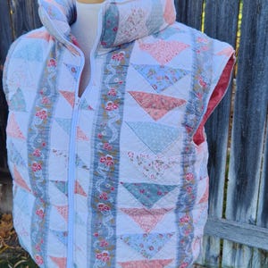 May include: A quilted vest with a high collar, featuring a patchwork design of triangles and squares in pastel colors. The vest has a full-length zipper and is displayed on a wooden form.