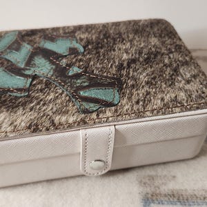 May include: A white jewelry box with a textured lid featuring a brown and gray pattern and turquoise accents. The box has a white snap closure and a rectangular shape, suitable for storing jewelry.