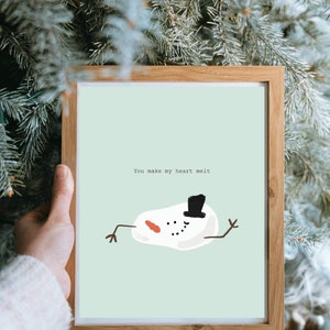 May include: A framed print featuring a light green background with a graphic of a melting snowman wearing a black top hat, orange carrot nose, and stick arms. The text "You make my heart melt" is printed in black above the snowman illustration.