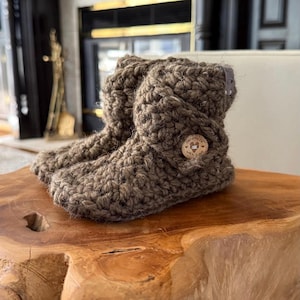 May include: A pair of handmade, brown crochet booties. The booties have a button closure on the side and are made of a thick, textured yarn. The booties are sitting on a wooden surface.