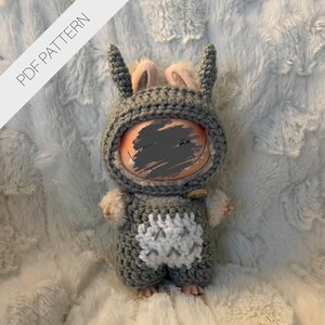 Puede incluir: Muñeca hecha a mano con un disfraz de conejito de ganchillo gris con un diseño de vientre blanco. La muñeca tiene un cierre de botón y una capucha con orejas de conejo. El fondo es una tela suave y blanca con textura. El texto "PDF PATTERN" está en la esquina superior izquierda.