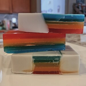 Può includere: Una pila di tre saponette a strati arcobaleno. La saponetta superiore è bianca con una striscia blu, arancione e gialla. La saponetta centrale è rossa, arancione, gialla, verde e blu. La saponetta inferiore è bianca con una striscia verde, arancione e rossa.