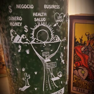 May include: Green glass candle with white text and illustrations. The text reads "Abre Camino", "Road Opener", "Negocio", "Business", "Dinero", "Money", "Health", "Salud", "Love", "Amor", "Exito", and "Success". The illustration depicts a road leading to a mountain with a sun behind it.