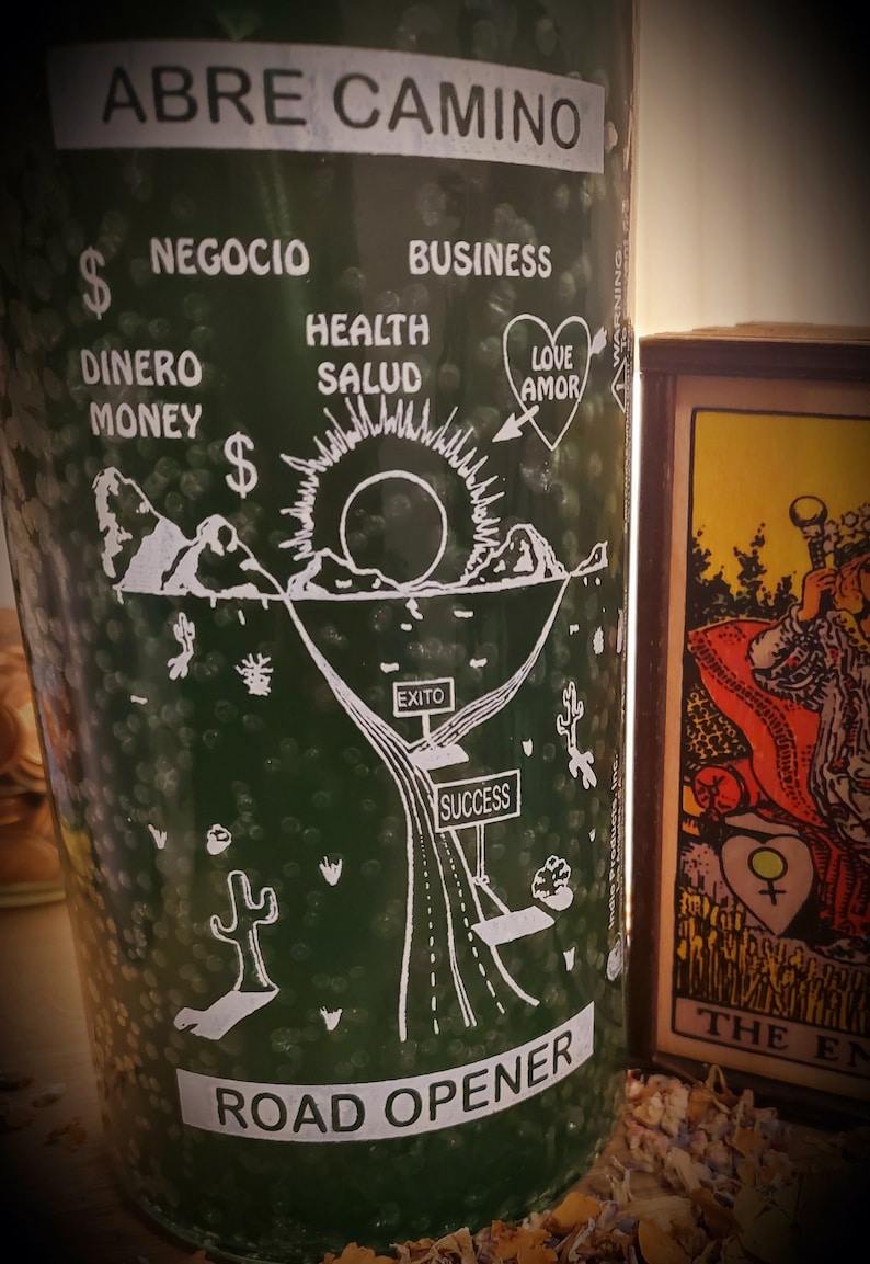 May include: Green glass candle with white text and illustrations. The text reads "Abre Camino", "Road Opener", "Negocio", "Business", "Dinero", "Money", "Health", "Salud", "Love", "Amor", "Exito", and "Success". The illustration depicts a road leading to a mountain with a sun behind it.