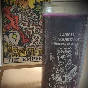 May include: A purple candle with a white image of a king holding a sword. The candle has the text "JUAN EL CONQUISTADOR BENDICION AL HOGAR" and "JOHN THE CONQUEROR HOUSE BLESSING CANDLE". The candle is next to a tarot card with the image of a queen on a throne. The card is labeled "THE EMPRESS".