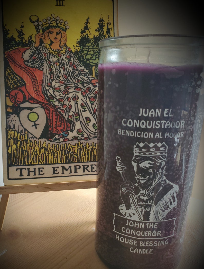 May include: A purple candle with a white image of a king holding a sword. The candle has the text "JUAN EL CONQUISTADOR BENDICION AL HOGAR" and "JOHN THE CONQUEROR HOUSE BLESSING CANDLE". The candle is next to a tarot card with the image of a queen on a throne. The card is labeled "THE EMPRESS".