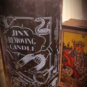 May include: A black glass candle jar with a white label that says "Jinx Removing Candle". The label also features various symbols including a horseshoe, a heart with an arrow, a snake, a dice, a cat, and a hand. The candle is sitting on a table with a tarot card that says "The Empress" in the background.
