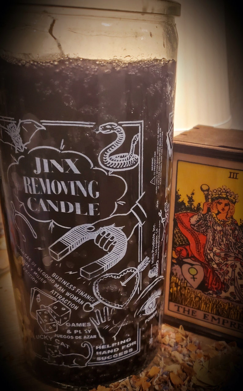 May include: A black glass candle jar with a white label that says "Jinx Removing Candle". The label also features various symbols including a horseshoe, a heart with an arrow, a snake, a dice, a cat, and a hand. The candle is sitting on a table with a tarot card that says "The Empress" in the background.