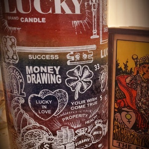 May include: A rainbow-colored glass candle with the words "LUCKY BRAND CANDLE" at the top. The candle has various symbols and words printed on it, including "MONEY DRAWING", "LUCKY IN LOVE", "YOUR WISH COME TRUE", "SUCCESS", "PROPERITY", "HEALTH", "FINANCE", and "33". The candle is sitting on a wooden surface with dried flowers.
