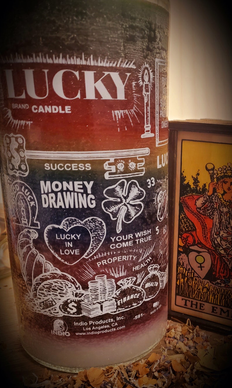 May include: A rainbow-colored glass candle with the words "LUCKY BRAND CANDLE" at the top. The candle has various symbols and words printed on it, including "MONEY DRAWING", "LUCKY IN LOVE", "YOUR WISH COME TRUE", "SUCCESS", "PROPERITY", "HEALTH", "FINANCE", and "33". The candle is sitting on a wooden surface with dried flowers.