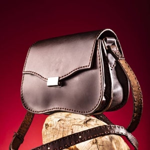 May include: A brown leather satchel bag with a silver buckle and a long strap. The bag is sitting on a piece of weathered wood.
