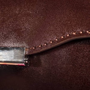 May include: Close-up of a brown leather bag with a silver metal clasp. The bag has a stitched seam around the edge.