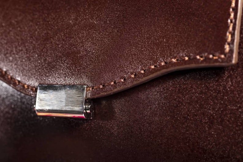 May include: Close-up of a brown leather bag with a silver metal clasp. The bag has a stitched seam around the edge.