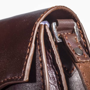 May include: Close-up of a brown leather bag with a silver buckle and stitching detail. The bag has a flap closure and a strap.