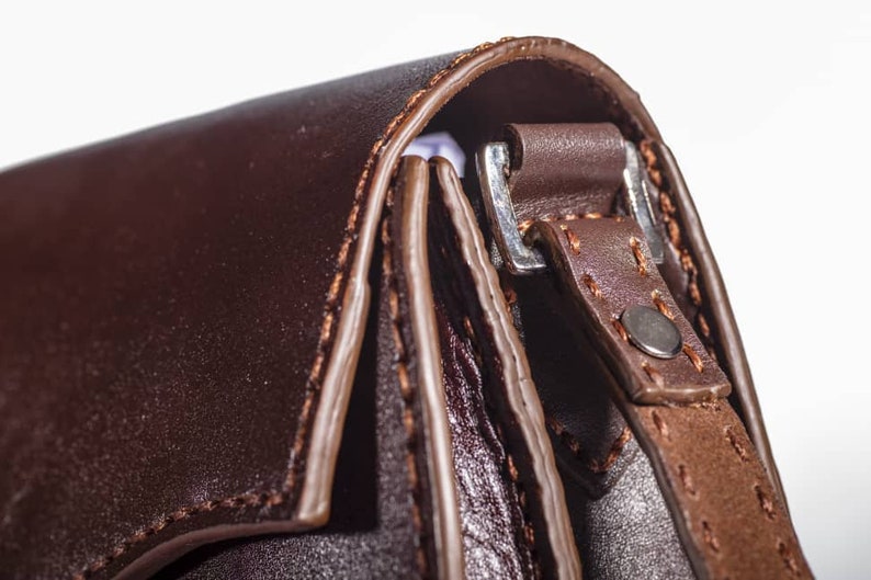 May include: Close-up of a brown leather bag with a silver buckle and stitching detail. The bag has a flap closure and a strap.